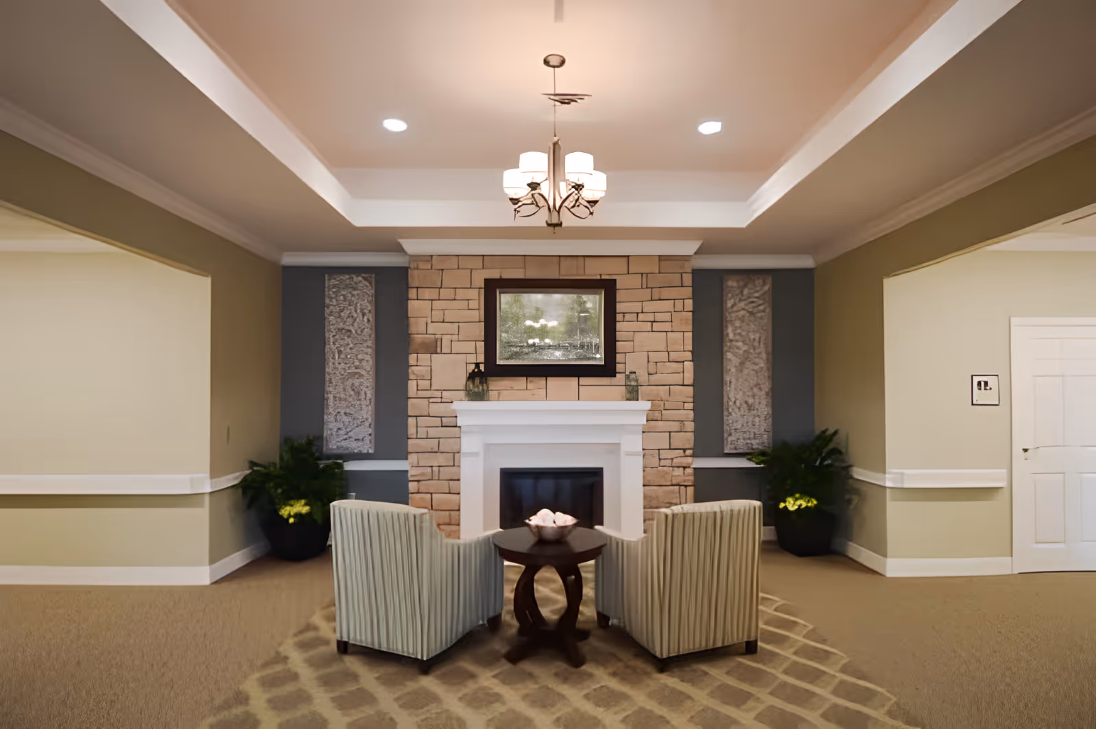 Two upholstered armchairs and a small table in a communal lounge facing a stone fireplace beneath a chandelier.