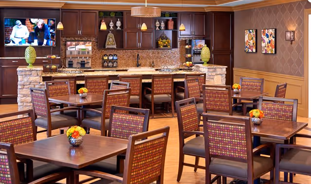 A dining area with multiple wooden tables and chairs featuring patterned upholstery. In the background, there is a kitchen counter with stone accents, dark wood cabinets, and pendant lighting. A television is mounted on the wall showing two people, and decorative items and artwork adorn the walls.