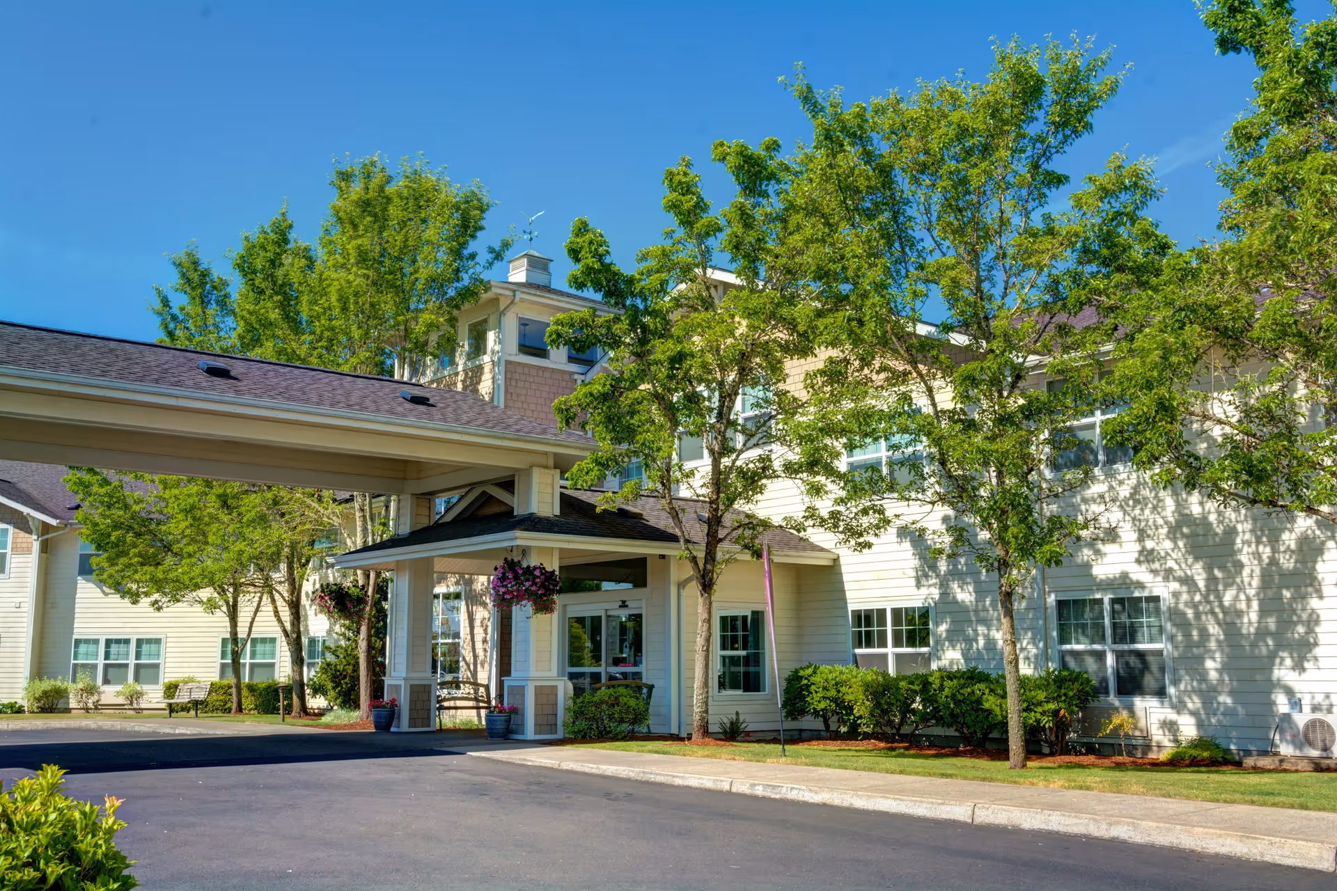 Exterior view of Vineyard Heights Assisted Living facility showing a covered entrance with hanging flower baskets, surrounded by green trees and shrubs under a clear blue sky.