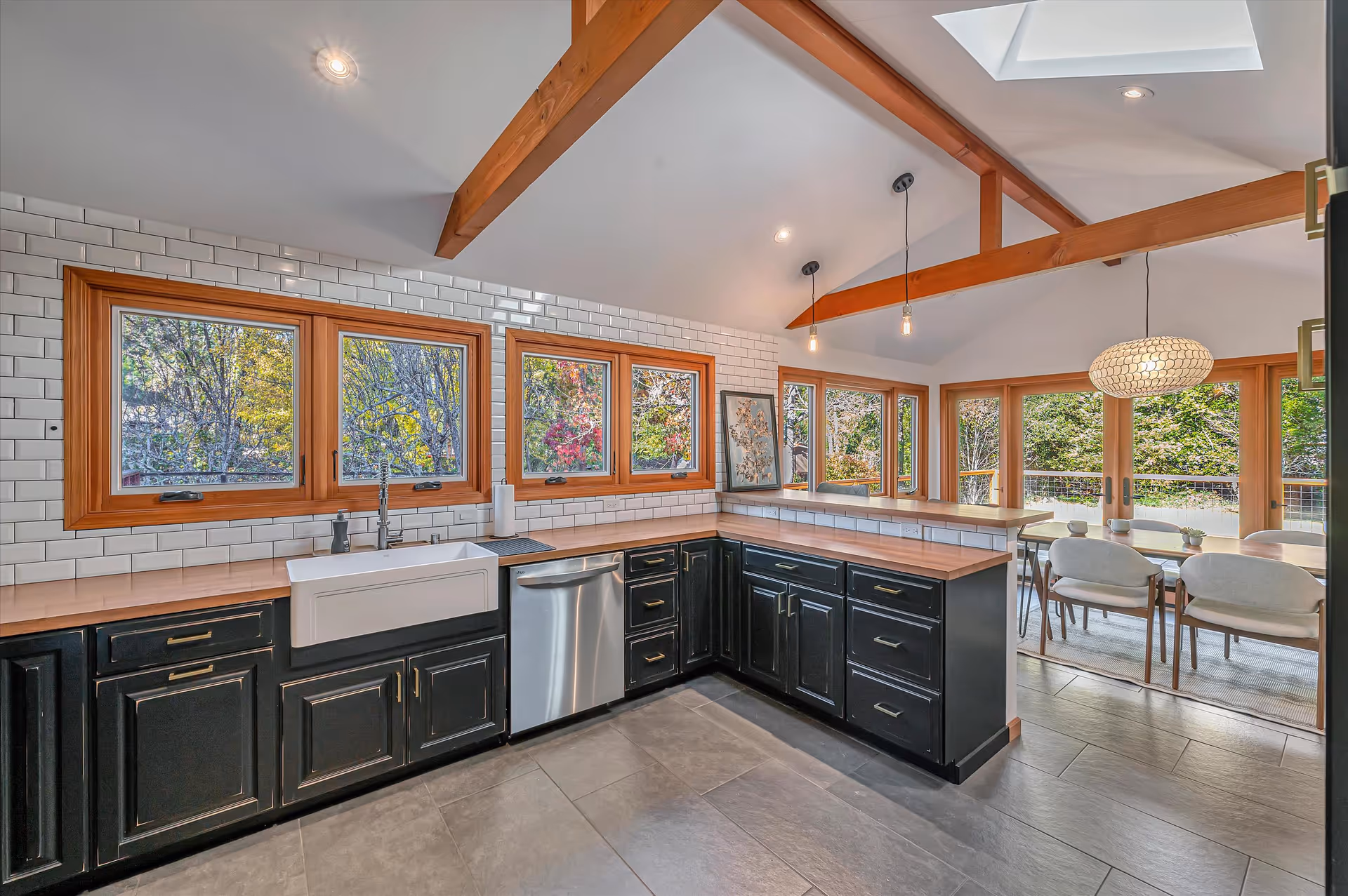 Modern kitchen with black cabinets, wooden countertops, white subway tile backsplash, farmhouse sink, stainless steel dishwasher, and multiple wooden-framed windows showing outdoor greenery. Adjacent dining area with a wooden table, white chairs, pendant lights, and large windows providing natural light.