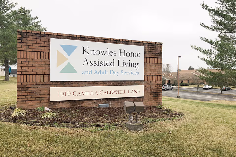 A brick sign outside displaying the name 'Knowles Home Assisted Living and Adult Day Services' with the address '1010 Camilla Caldwell Lane'. The sign is situated on a grassy area with trees and a parking lot with cars in the background.