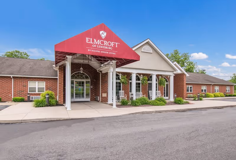 Front exterior view of a senior living facility named Elmcroft of Lewisburg with a red canopy entrance, white columns, hanging plants, and rocking chairs on the porch.