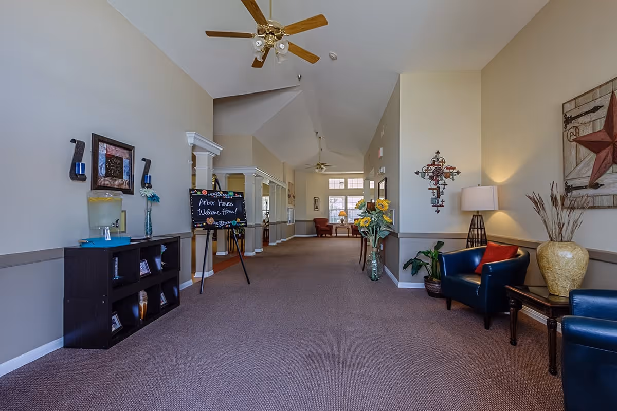 A spacious hallway in a senior living facility with beige walls and carpeted floor. The hallway features decorative elements including framed artwork, a large vase with sunflowers, a blackboard sign that reads 'Arbor House Welcome Home!', and comfortable seating with blue armchairs and a side table with a lamp. Ceiling fans are mounted on the high ceiling, and natural light comes through windows at the far end.