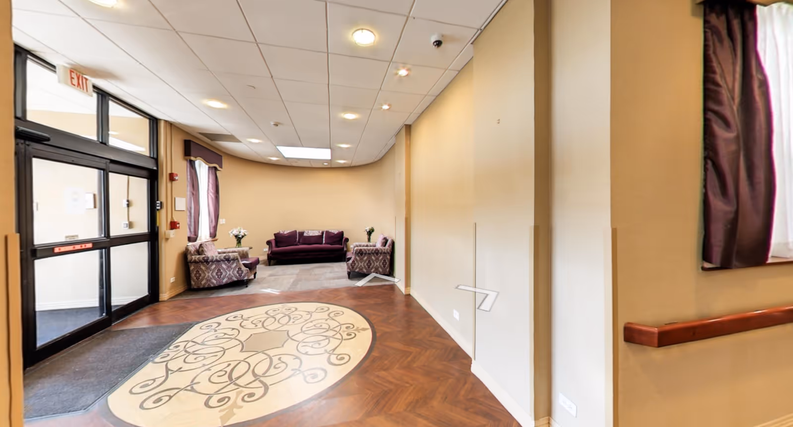 Interior view of a senior living facility lobby area with a decorative circular floor design, a seating area with a purple sofa and two patterned armchairs, beige walls, ceiling lights, and a glass exit door.