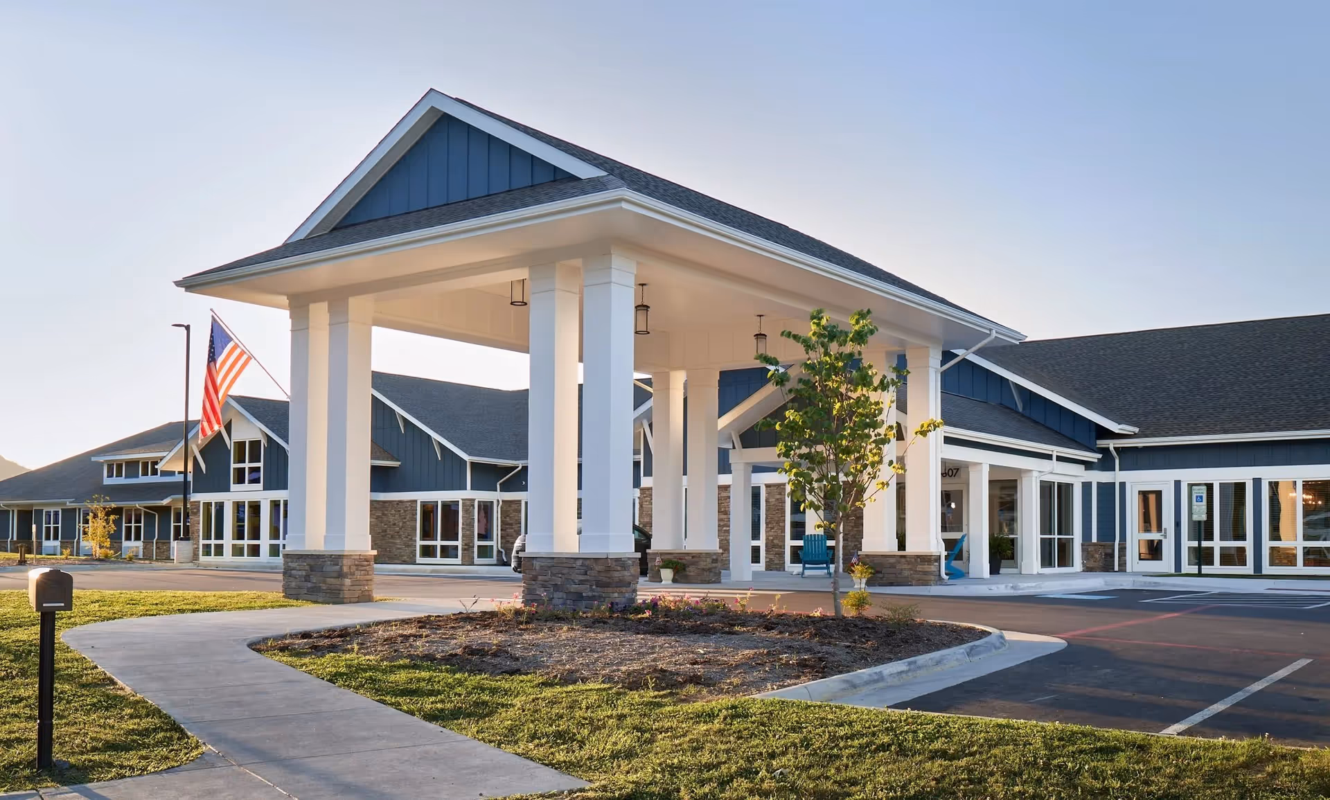 Exterior view of English Meadows Harrisonburg Campus building with a covered entrance supported by white pillars, an American flag on a flagpole, a small tree, and a paved driveway and walkway in the foreground under a clear sky.