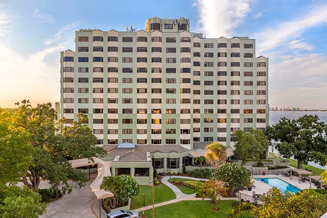 Exterior view of a multi-story senior living facility building with balconies, surrounded by trees and greenery. In front of the building, there is a well-maintained garden area with pathways, a swimming pool, and outdoor seating. The sky is partly cloudy with soft sunlight.