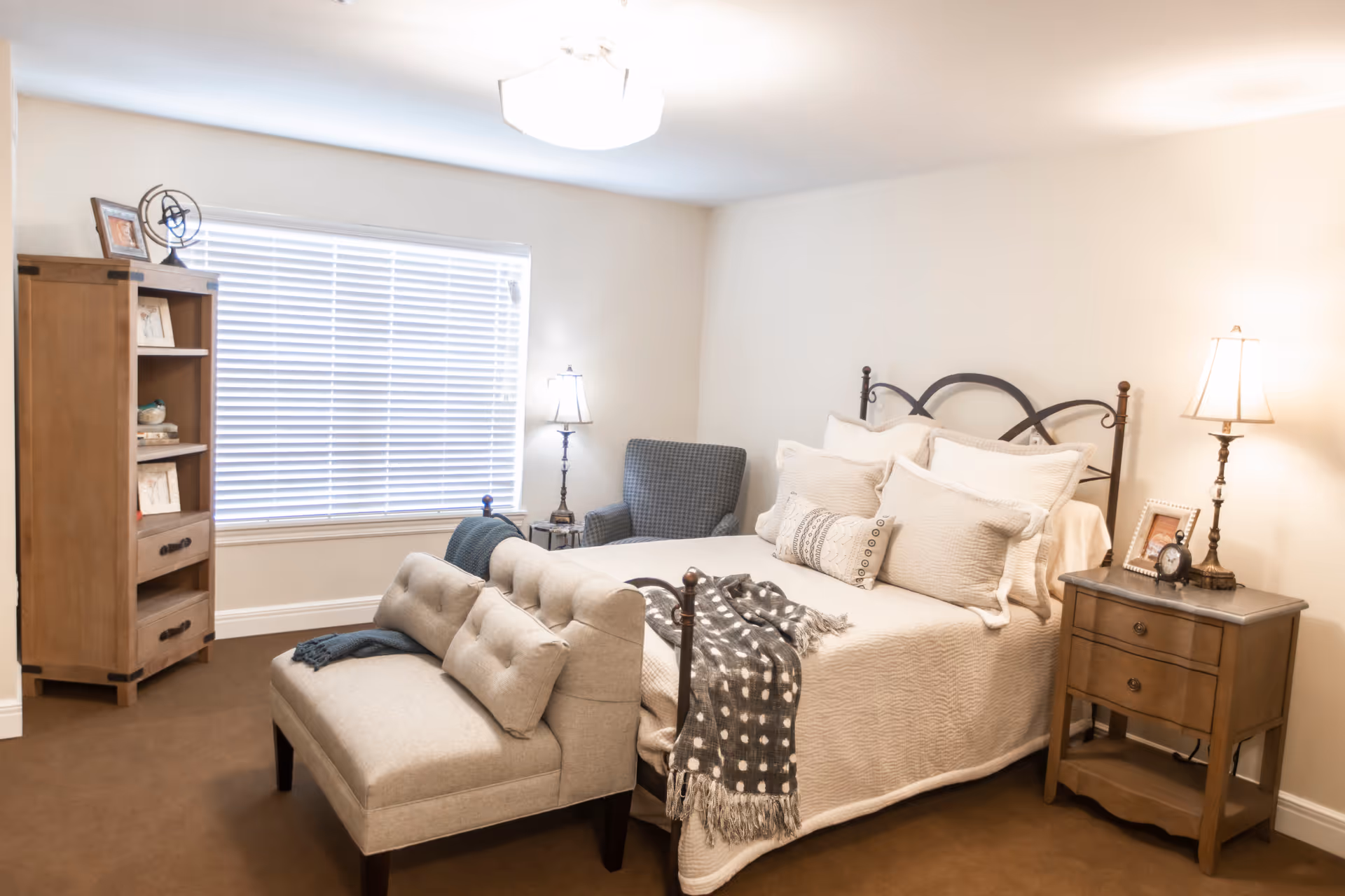 A cozy bedroom with a neatly made bed featuring multiple pillows and a throw blanket. There is a cushioned bench at the foot of the bed, a wooden nightstand with a lamp and a framed photo, a comfortable armchair in the corner, and a wooden bookshelf near a window with blinds.