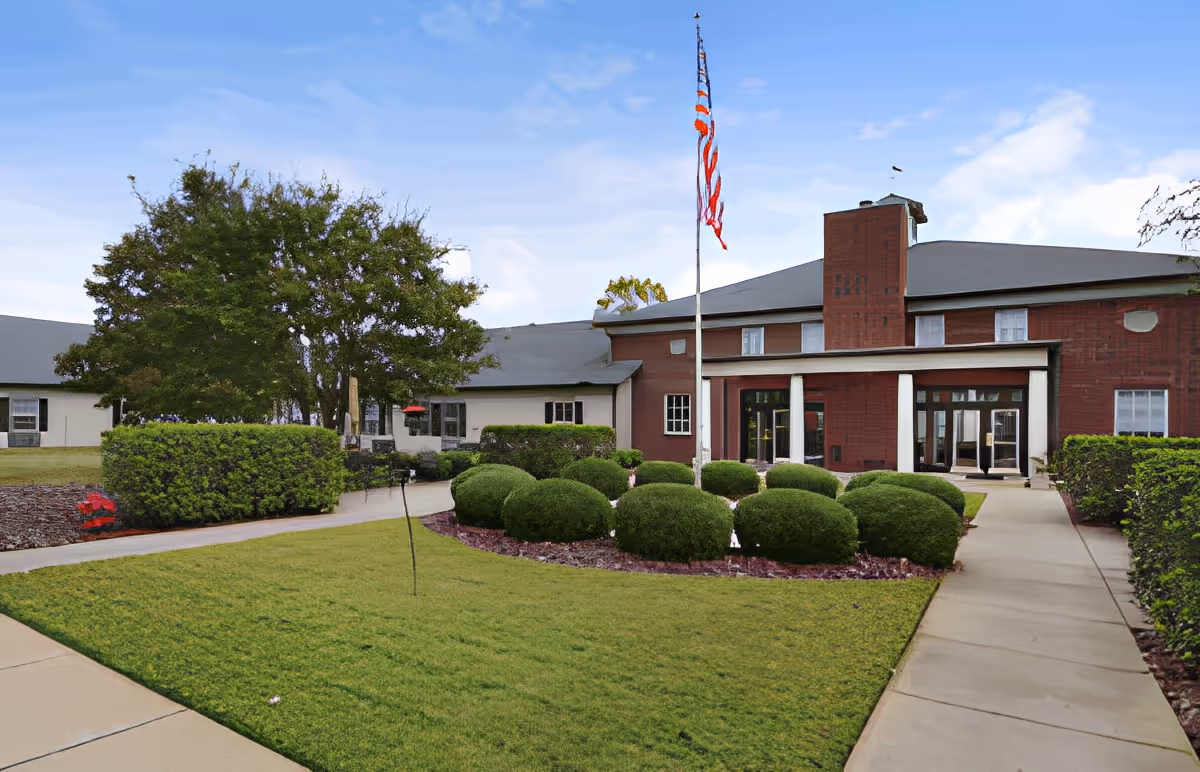 Front entrance of a red brick senior living building with an American flag, manicured bushes, and paved walkways.