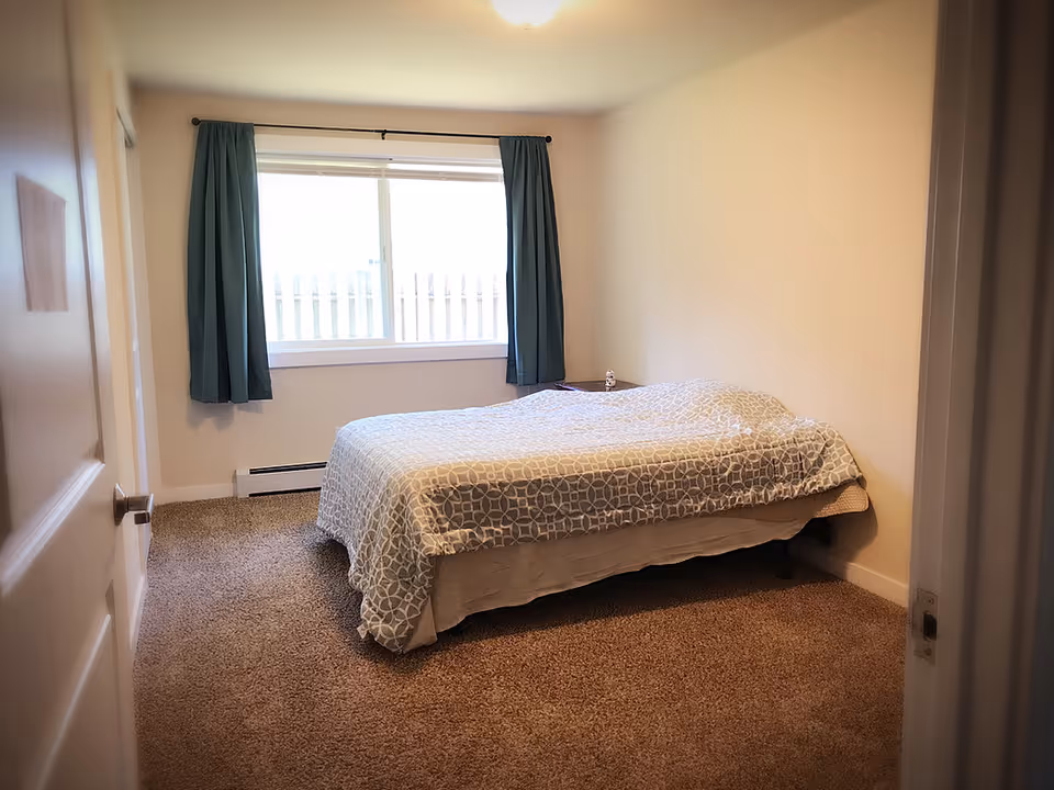 A simple bedroom with a made bed, teal curtains over a window, and beige carpeted floor.