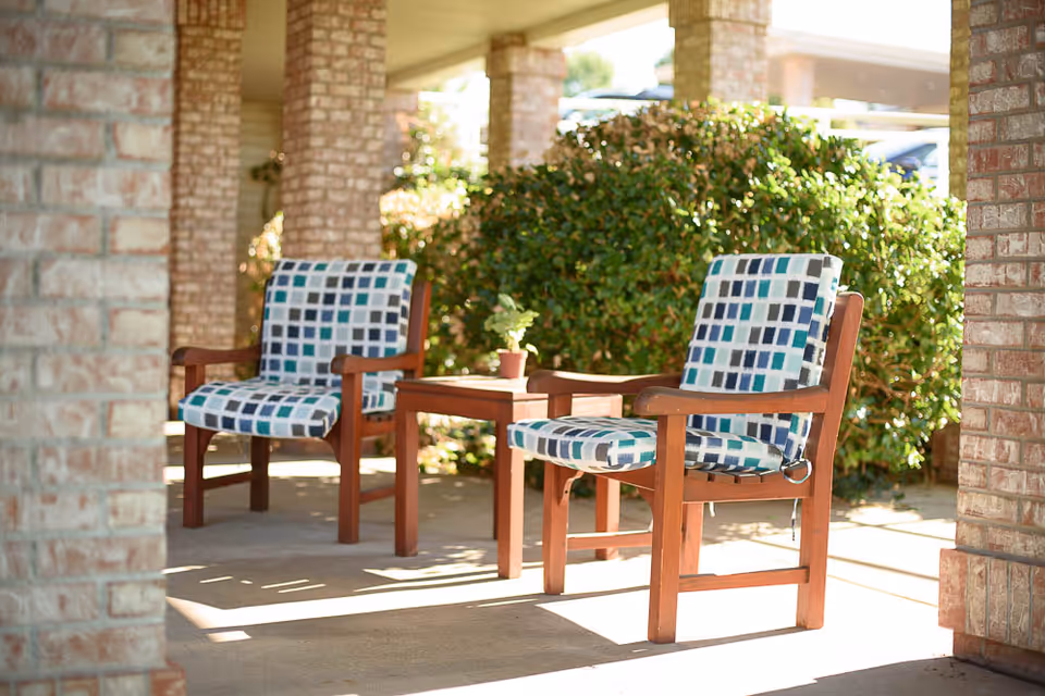 Two cushioned wooden chairs and a small table on a covered outdoor patio with brick columns and greenery.