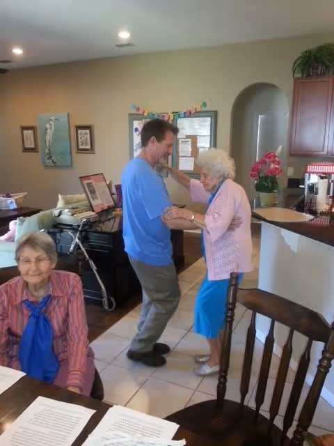 An elderly woman in a pink cardigan and blue skirt is dancing with a man in a blue shirt inside a senior living facility. Another elderly woman wearing a striped shirt and blue scarf is sitting at a table with papers in front of her. The room has light-colored tile flooring, wooden furniture, and some decorations including framed pictures and a plant on a counter.