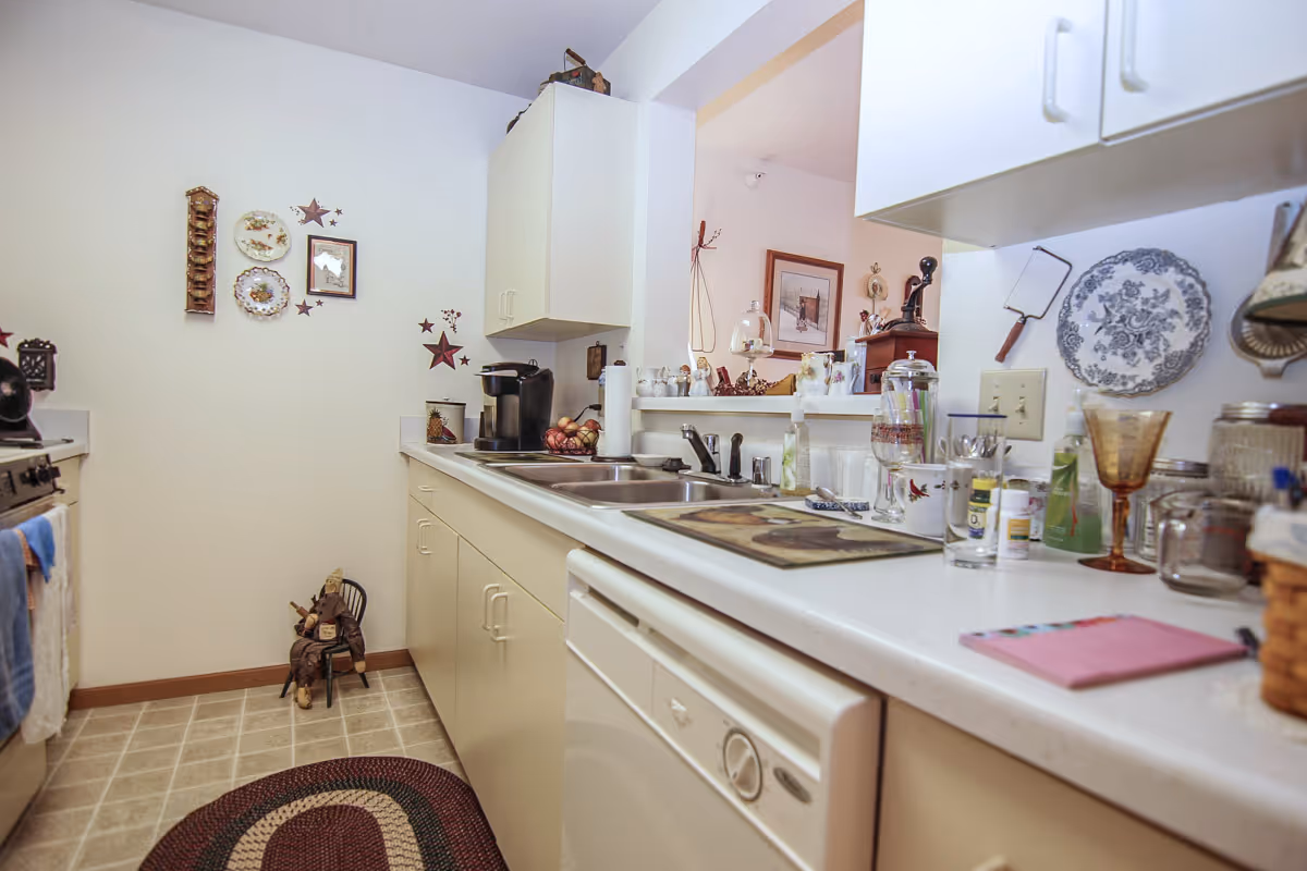A compact kitchen with white cabinets and countertops. The kitchen features a double sink, a dishwasher, a coffee maker, and various kitchen items such as glasses, bottles, and decorative plates on the wall. There is a small rug on the tiled floor and a decorative figure sitting on a chair in the corner. The kitchen opens to another room with framed pictures and additional decor visible.
