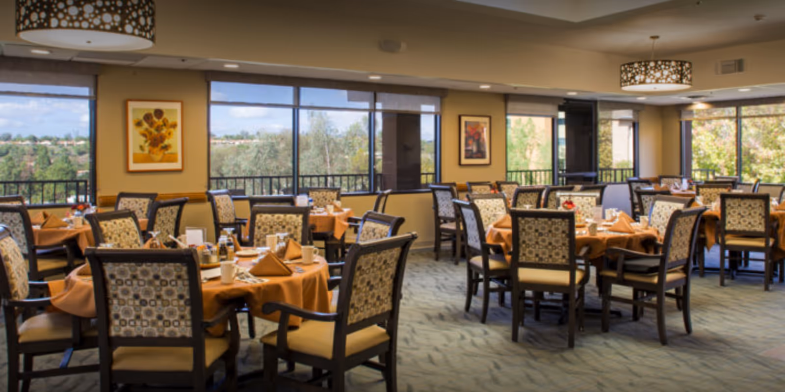 A spacious dining room with multiple round tables covered with orange tablecloths, set with napkins, cups, and condiments. The chairs have patterned upholstery and wooden frames. Large windows provide a view of trees and a partly cloudy sky outside. The room is well-lit with ceiling lights and decorated with framed artwork on the walls.