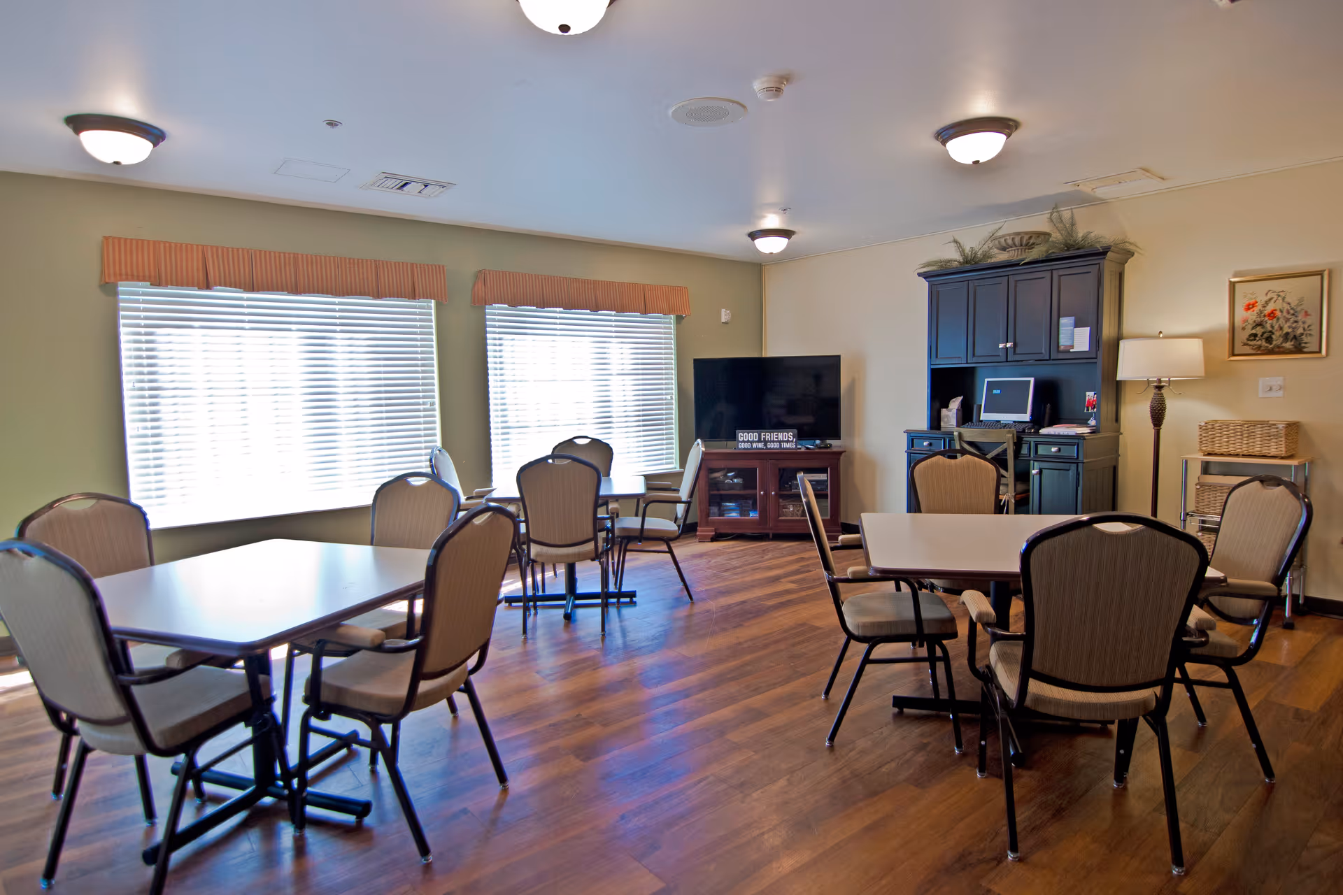 Communal dining/activity room with several tables and chairs, a TV and storage cabinet, and large windows with blinds.