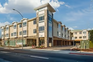 Exterior view of a modern multi-story senior living facility building with beige and white walls, large windows, and a covered entrance. The building is situated along a street with a sidewalk and street lamps under a partly cloudy sky.