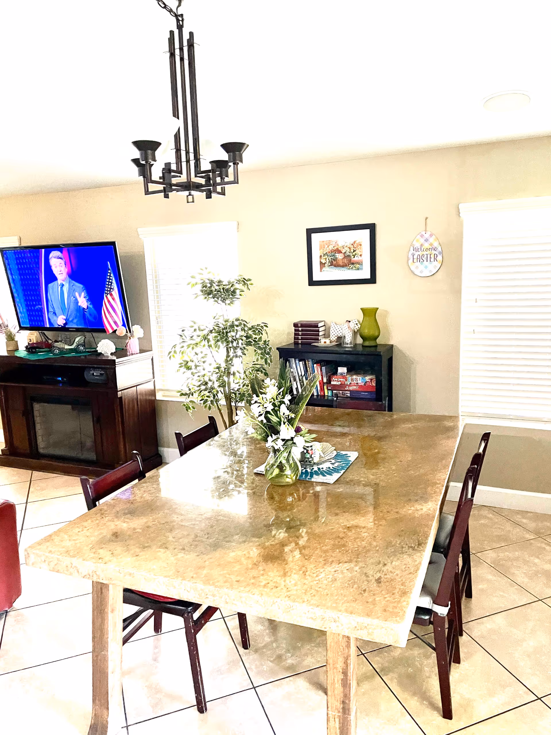 A bright dining room with a large rectangular stone table surrounded by wooden chairs. On the table is a vase with white flowers and some decorative items. In the background, there is a TV on a wooden stand, a potted plant, a bookshelf with books and games, and wall decorations including a framed picture and a sign that says 'Welcome Easter'. The room has tiled floors and windows with white blinds.