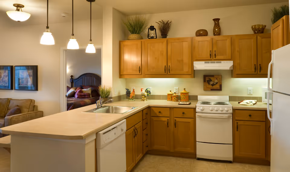 Open kitchen with wooden cabinets, white appliances, a peninsula with sink and pendant lights, and a view into a living area and bedroom.
