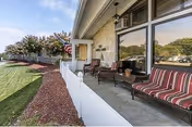 Covered front porch of a senior living facility featuring cushioned outdoor chairs, potted plants, a low white fence, an American flag, and an adjacent lawn.
