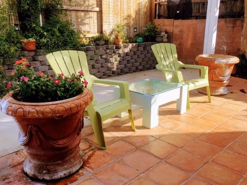 Outdoor patio area with two green plastic chairs and a small white table with a glass top between them. There are large decorative terracotta planters with flowering plants, a stone retaining wall with greenery, and a wooden fence in the background.