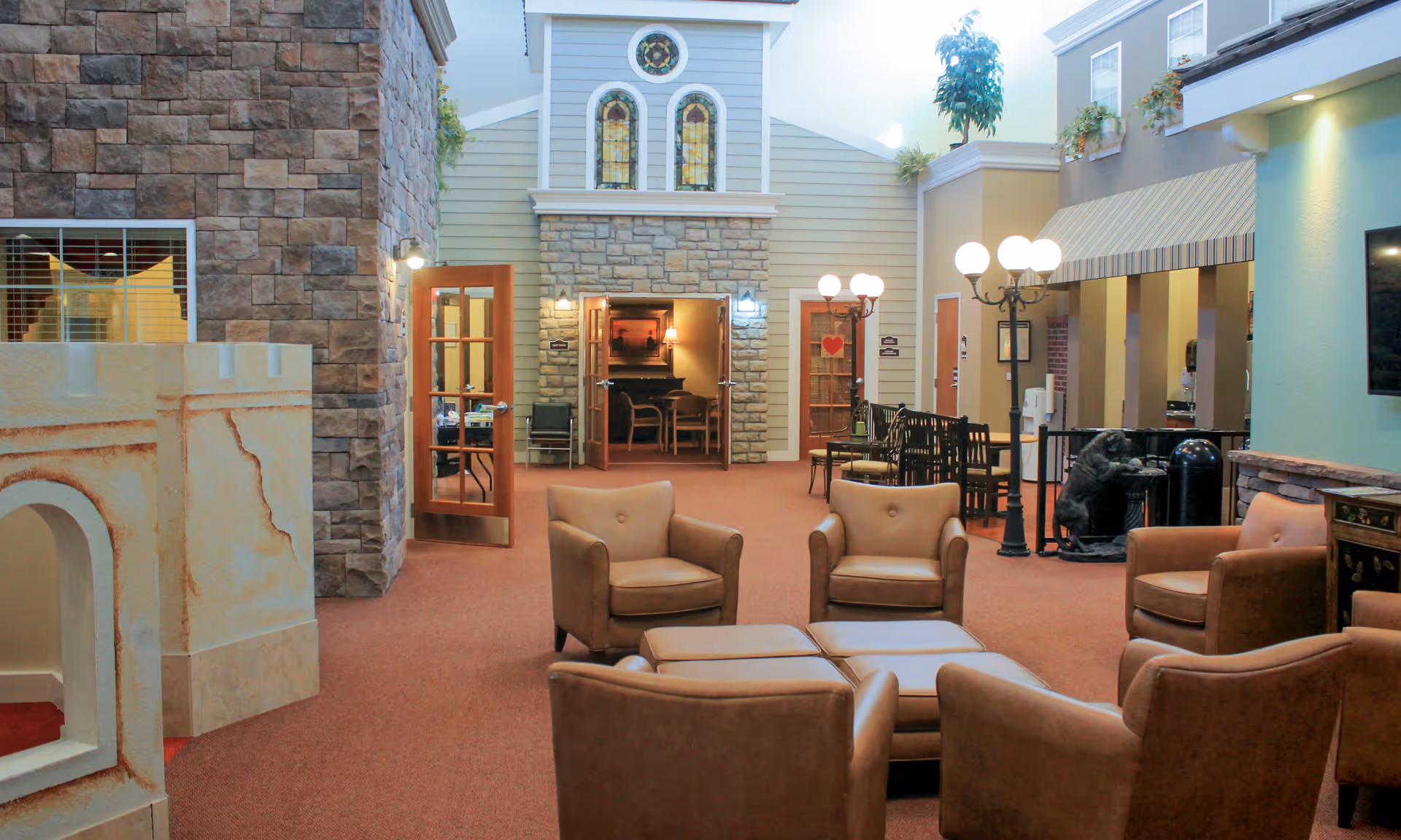 A cozy indoor common area with four brown leather armchairs arranged around two ottomans on a carpeted floor. The room features stone and wood panel walls, decorative stained glass windows, and multiple doorways leading to other rooms. There are vintage-style street lamps with globe lights, potted plants on ledges, and a bear statue drinking from a fountain near a small dining area with tables and chairs.