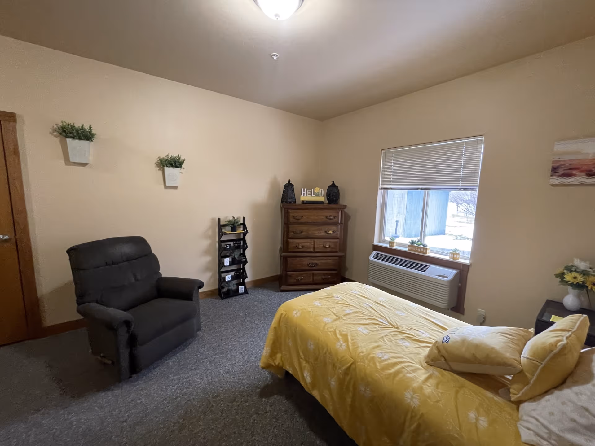 A cozy bedroom with beige walls and gray carpet. The room features a bed with yellow bedding and pillows, a dark gray recliner chair, a wooden dresser with decorative items on top, a small black shelf with plants and framed pictures, and a window with blinds partially open letting in natural light. There are two small potted plants hanging on the wall and a small table with a vase of flowers next to the bed.