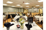 A dining room with multiple tables covered in white tablecloths, each set with black folded napkins, cups, and small flower arrangements. The room has wooden flooring, beige walls, and ceiling lights, with chairs arranged around the tables.