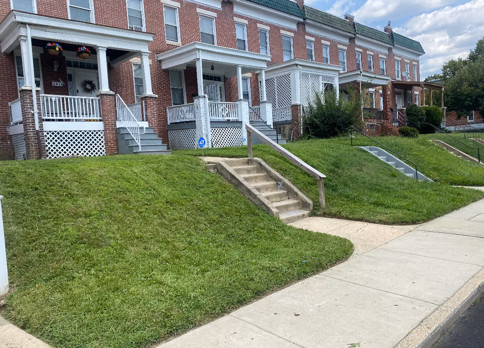 Row of brick townhouses with front porches, steps, and grassy lawns along a sidewalk.