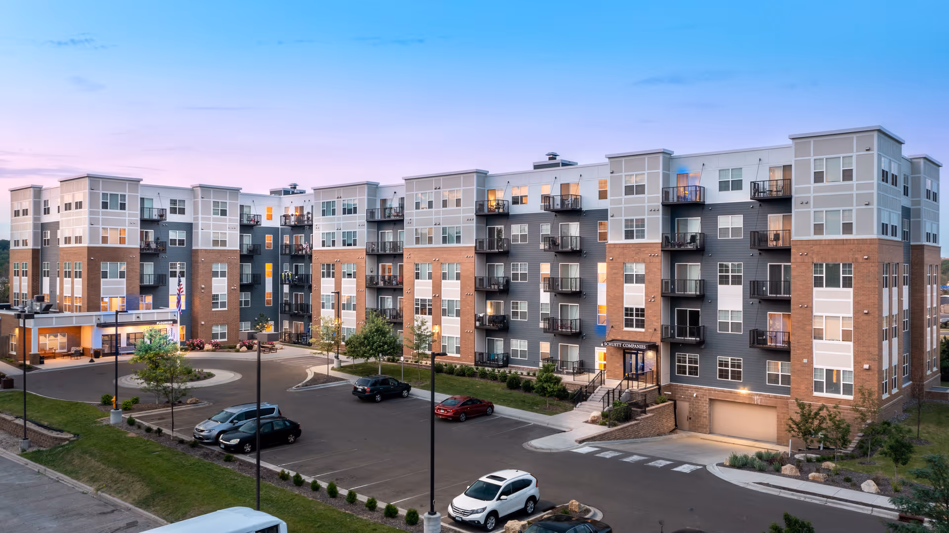 Exterior front view of a multi-story Flourish Senior Living apartment building with balconies, entrance canopy, and a parking lot at dusk.