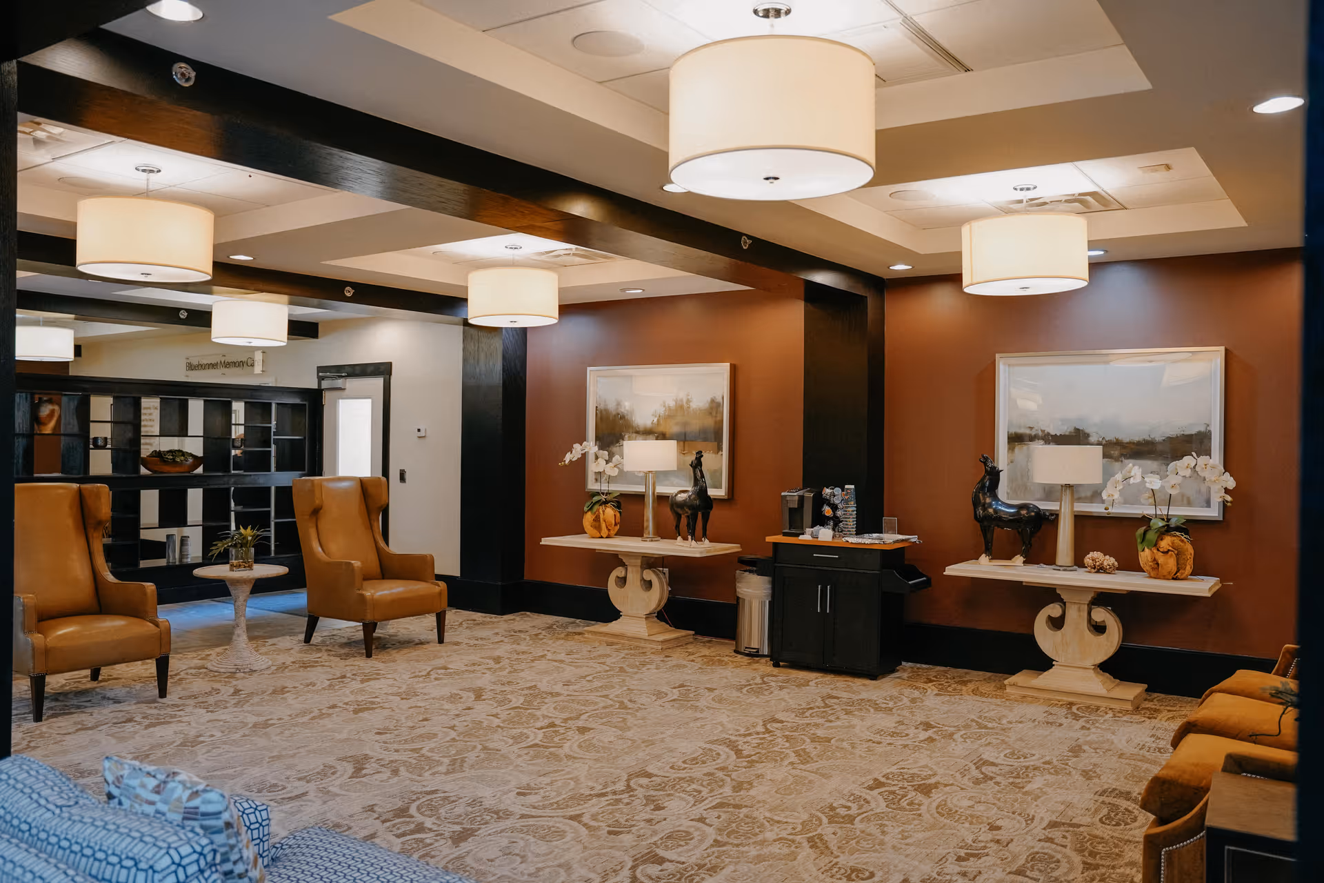 A spacious and well-lit common area with beige patterned carpet, brown leather armchairs, and a blue patterned sofa. The walls are painted in a warm brown tone with black trim, and there are two decorative tables with lamps, sculptures, and plants. Large ceiling lights provide soft illumination, and a black shelving unit is visible in the background.