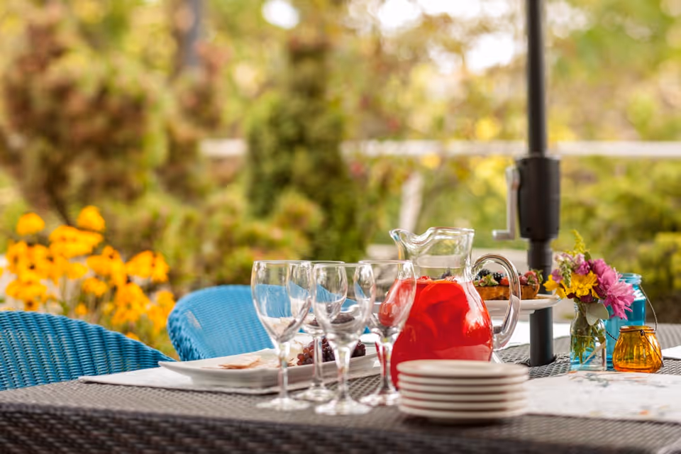 Outdoor patio table set with wine glasses, a pitcher of red beverage, stacked plates and small flower arrangements against a blurred garden background.