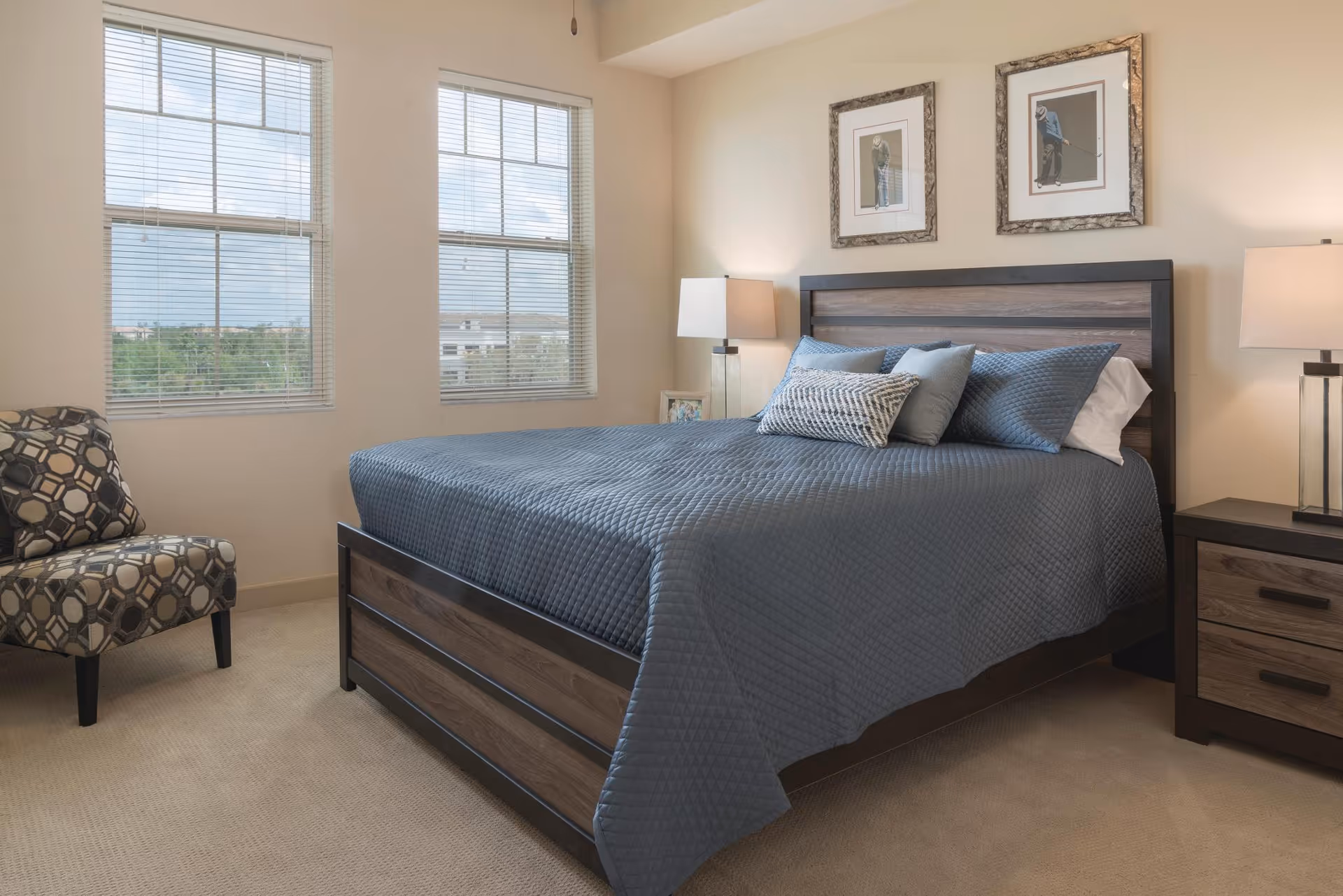 A bedroom with a large bed covered in a blue quilt and several pillows. There are two framed pictures hanging above the headboard. On either side of the bed are nightstands with modern lamps. To the left, there is a patterned armchair near two large windows with blinds, letting in natural light.