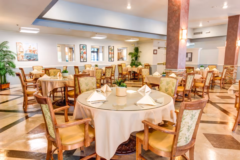 A bright and spacious dining room with multiple round tables covered with beige tablecloths and set with folded napkins, utensils, and small potted plants. The chairs have wooden frames with cushioned seats and backs featuring a leafy pattern. The room has polished tile floors with a geometric design, light-colored walls adorned with framed artwork, and several large plants adding greenery. Soft lighting and decorative columns enhance the welcoming atmosphere.
