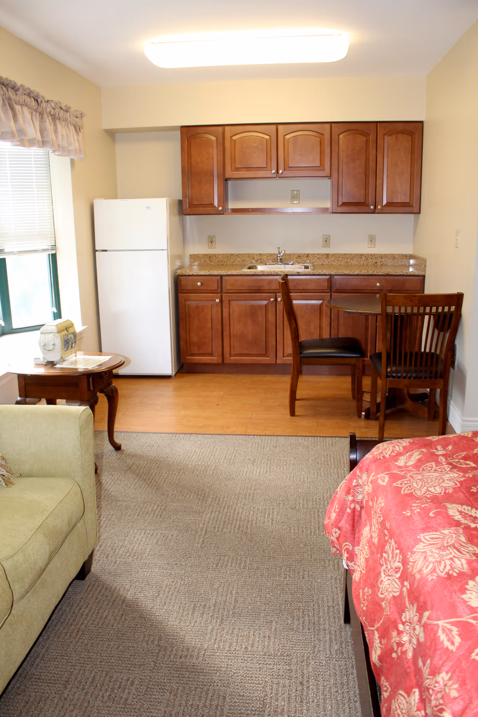 Studio-style room showing a kitchenette with wooden cabinets, a small dining table and chairs, a sofa and a bed.