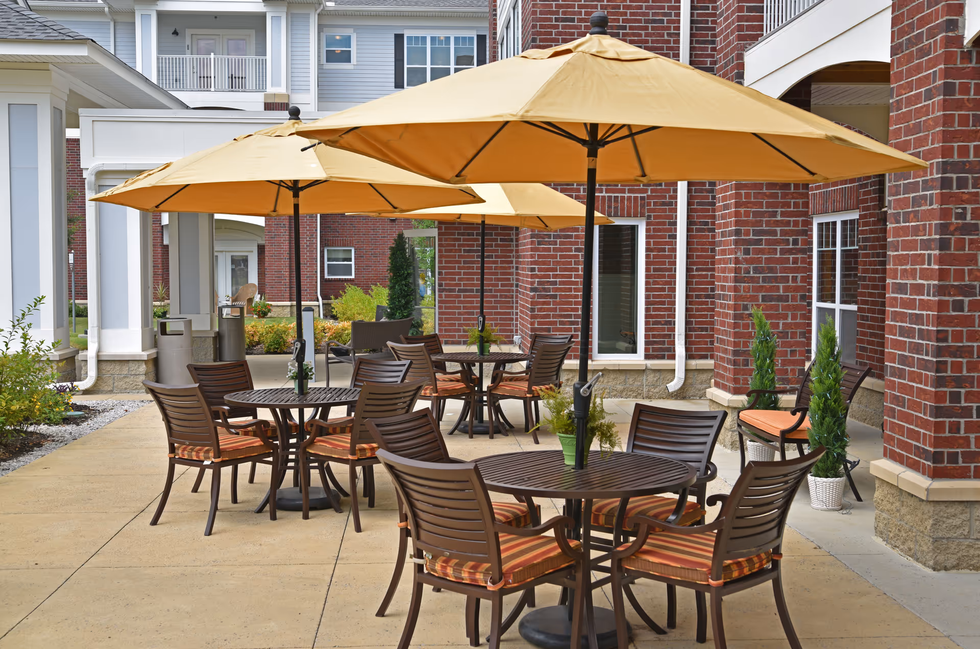 Outdoor patio area at an assisted living facility with round tables, brown chairs with striped cushions, and large yellow umbrellas providing shade. The patio is adjacent to a brick building with windows and some potted plants.