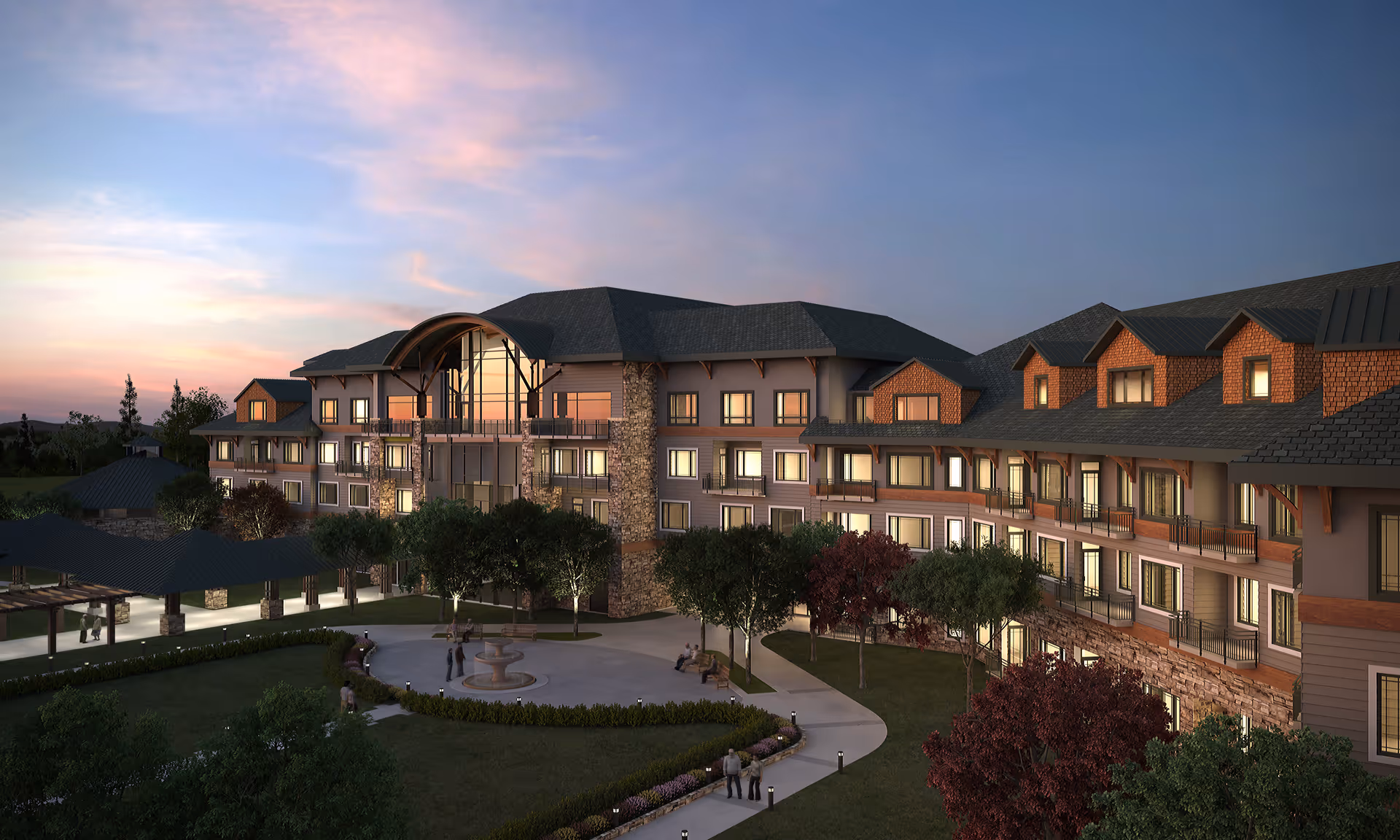 Exterior view of a large senior living facility building at sunset with multiple floors, balconies, and a landscaped courtyard featuring a fountain, trees, and walking paths. The sky is partly cloudy with soft pink and blue hues.