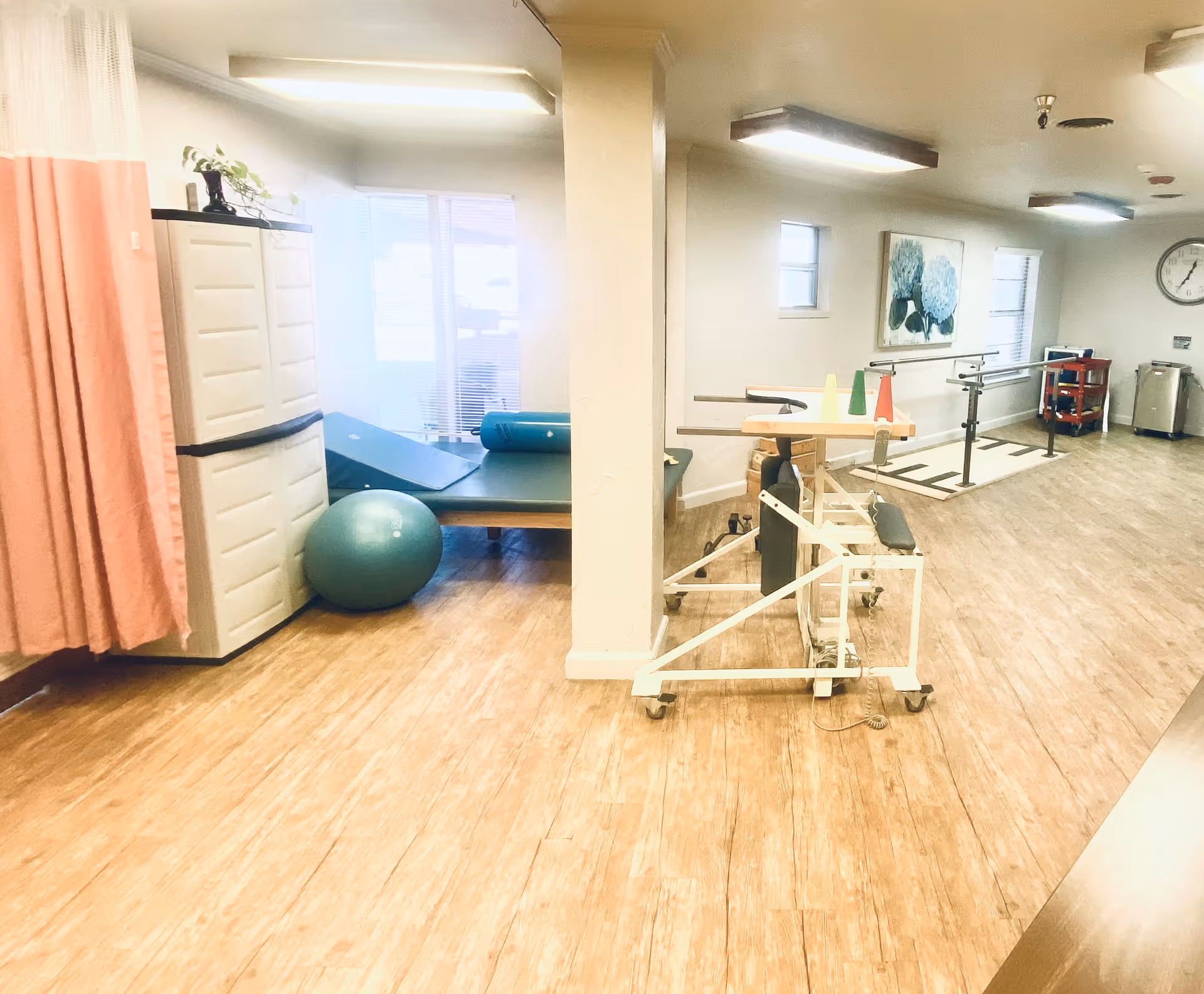 A rehabilitation room with wooden flooring, featuring parallel bars for walking exercises, a padded therapy table, an exercise ball, and a storage cabinet. The room is well-lit with ceiling lights and has windows with blinds. There is a clock on the wall and a painting of blue flowers.