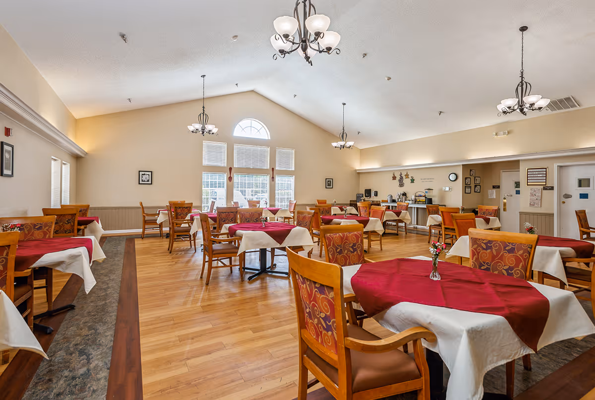 Spacious dining room with multiple tables dressed in white and red tablecloths, wooden chairs, chandeliers, and large arched windows.