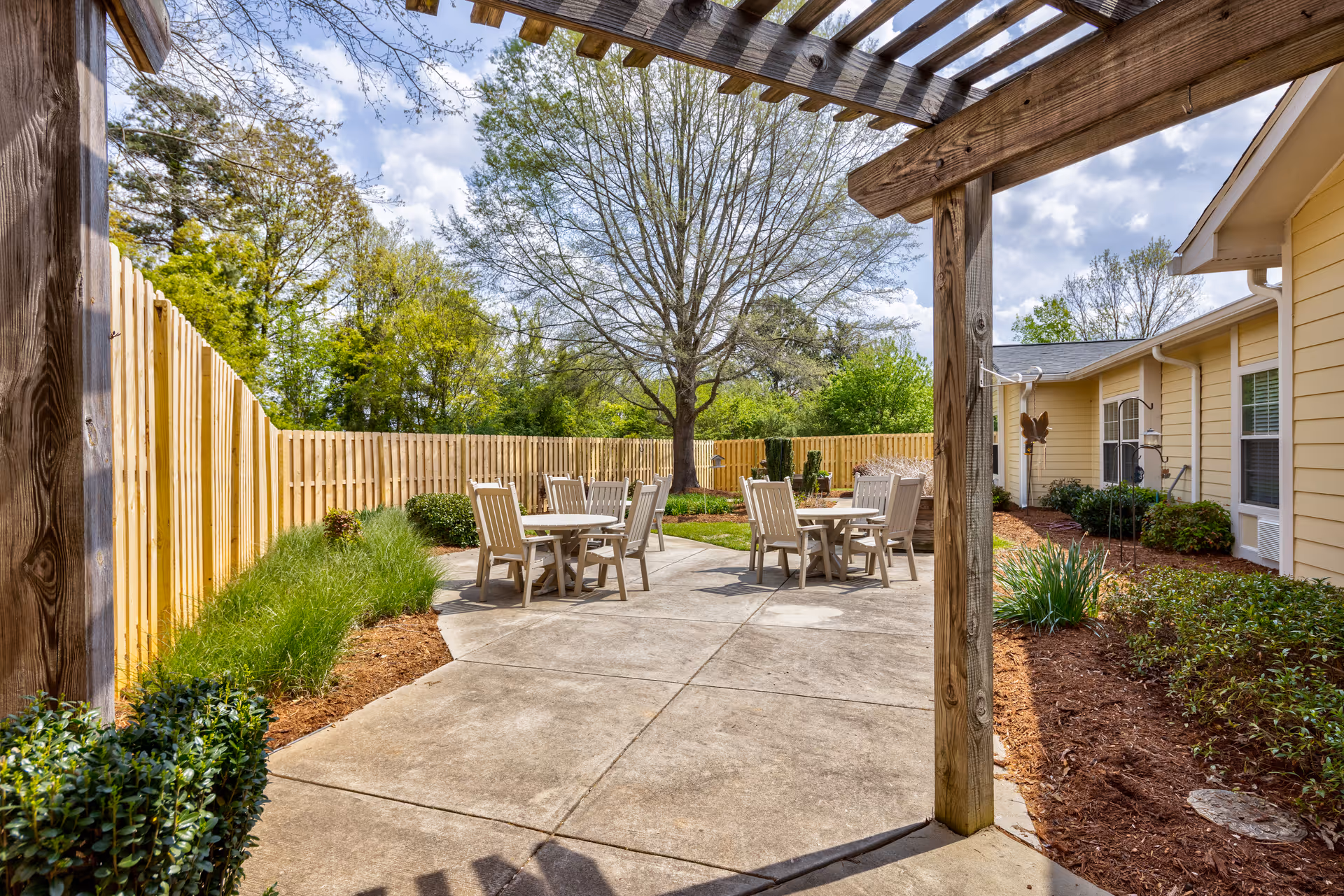 Sunny outdoor courtyard with round tables and chairs beneath a wooden pergola, surrounded by landscaping and a wooden fence beside a single-story building.