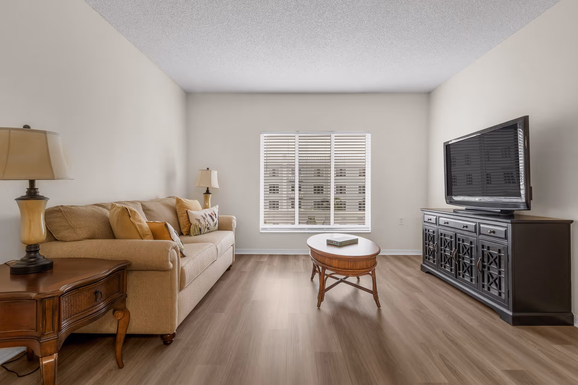 Living room with a beige sofa, wooden coffee table, large TV on a dark console, and a window with blinds.