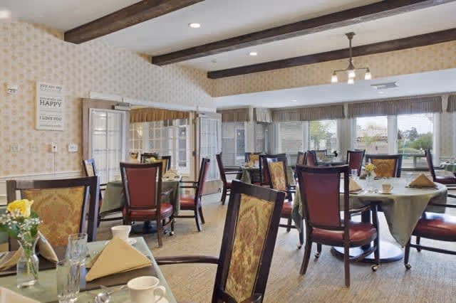 A bright dining room with multiple round tables covered with green tablecloths, each set with napkins, cups, glasses, and small flower arrangements. The room has large windows with a view outside, wooden beams on the ceiling, and patterned wallpaper.