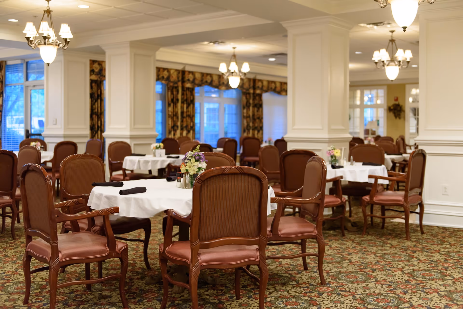 A spacious dining room with multiple round tables covered with white tablecloths and set with floral centerpieces and black napkins. The room features wooden chairs with cushioned seats and backs, patterned carpet, large white columns, and elegant chandeliers hanging from the ceiling. Windows with floral curtains allow natural light to enter the room.