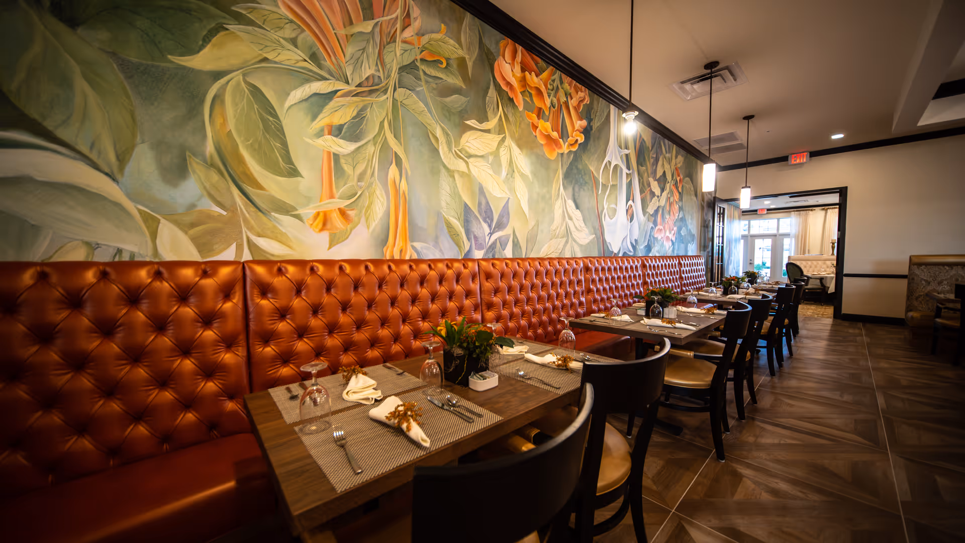 A dining area with a long tufted brown leather bench along the wall, several wooden tables set with placemats, napkins, silverware, and upside-down wine glasses. The wall behind the bench features a large, colorful mural of flowers and leaves. The floor has a dark wood pattern, and pendant lights hang from the ceiling.