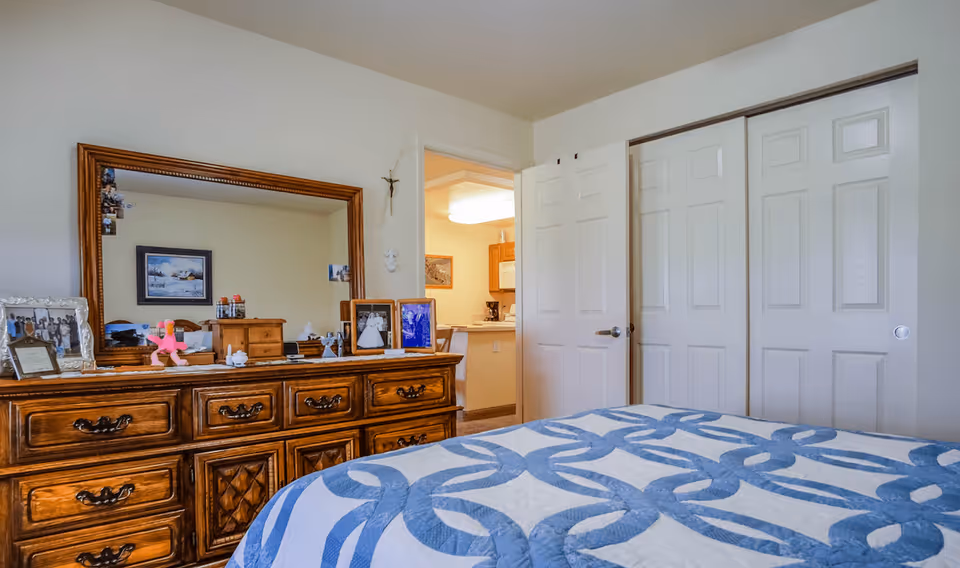 A bedroom in Devonshire Retirement Village featuring a wooden dresser with a large mirror above it. The dresser holds framed photos and small decorative items. A bed with a blue and white patterned quilt is partially visible in the foreground. The room has white walls, a door leading to another room, and a closet with sliding doors.