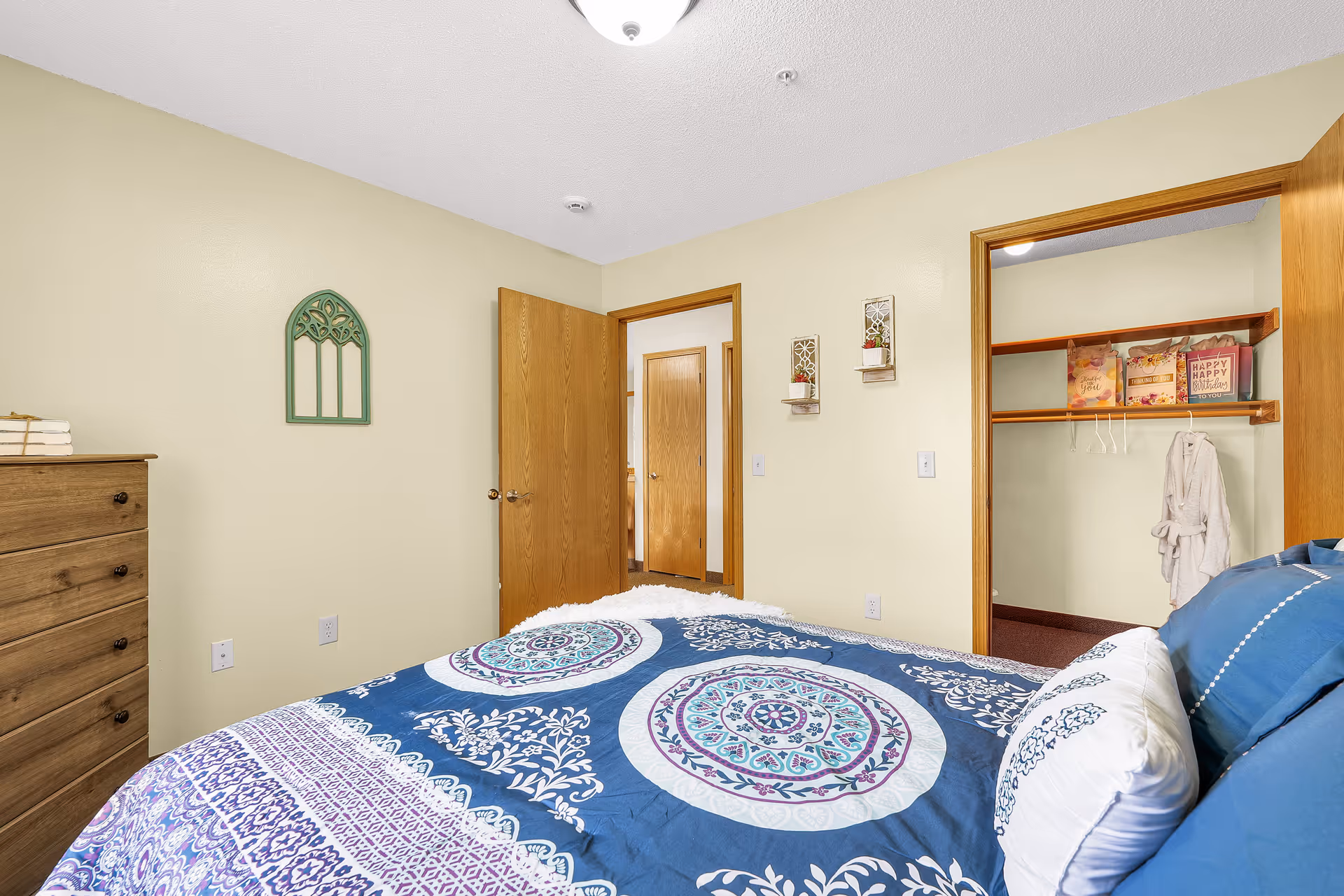 A bedroom in an assisted living facility featuring a bed with blue and white patterned bedding, a wooden dresser, and an open closet with shelves and hanging space. The walls are painted light yellow, and there are two wooden doors, one leading to a hallway and the other to the closet.