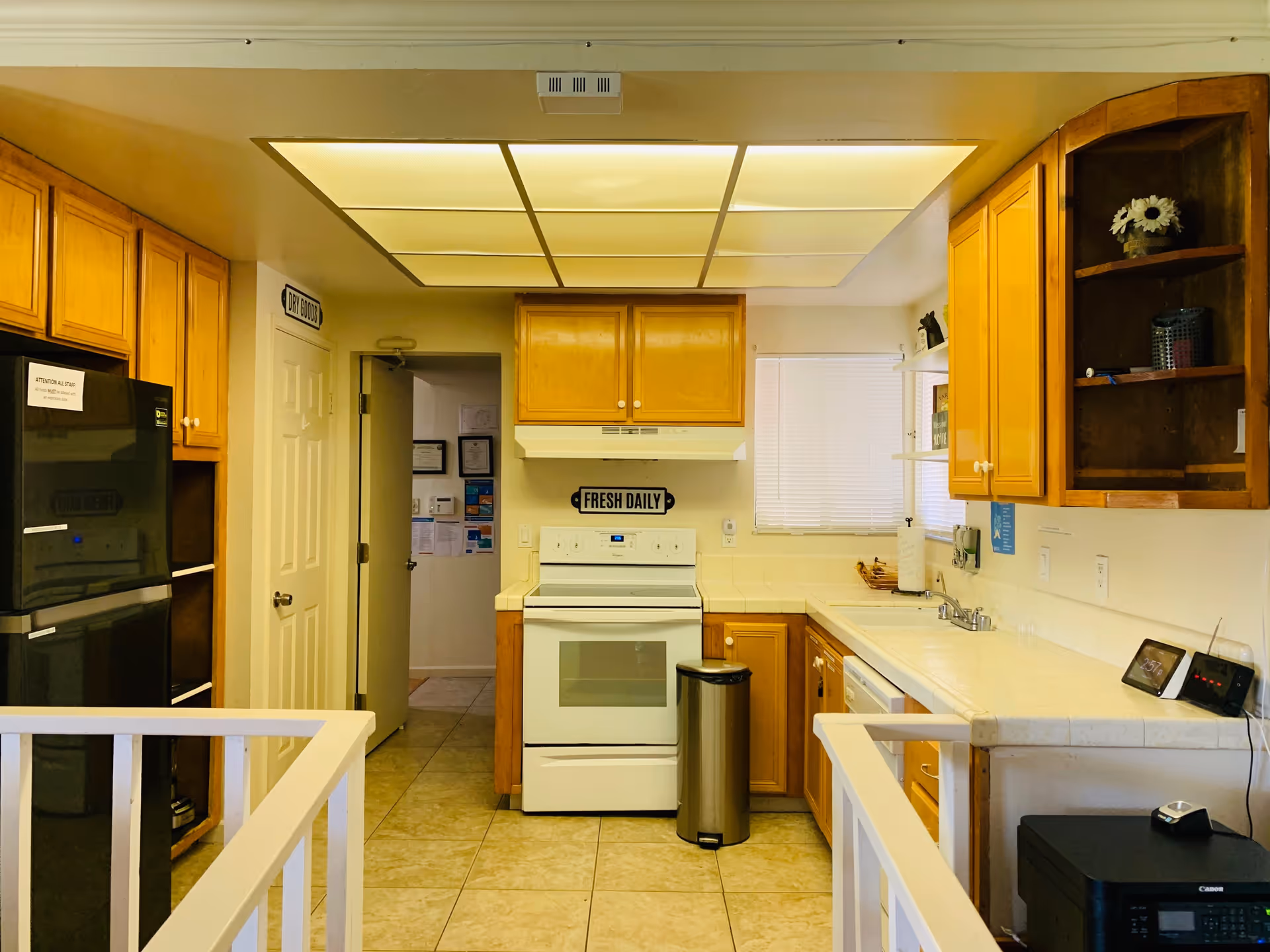 A kitchen with wooden cabinets, a white stove with a 'Fresh Daily' sign above it, a stainless steel trash can, a black refrigerator with a sign on it, a white tiled countertop with a sink, and various small items including a digital clock and a printer.