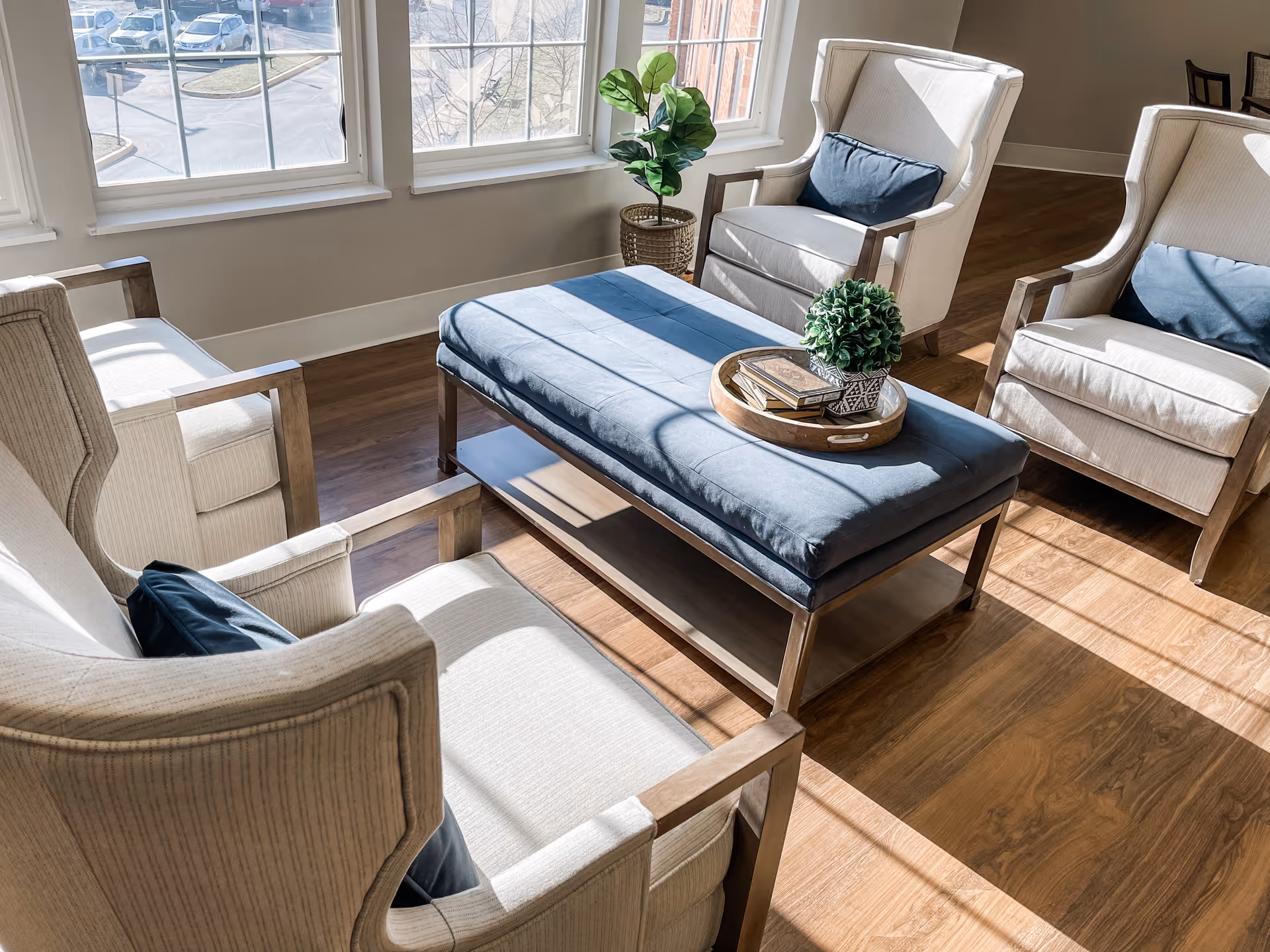 Sunlit seating area with four upholstered armchairs arranged around a blue upholstered ottoman on a wood floor by large windows.