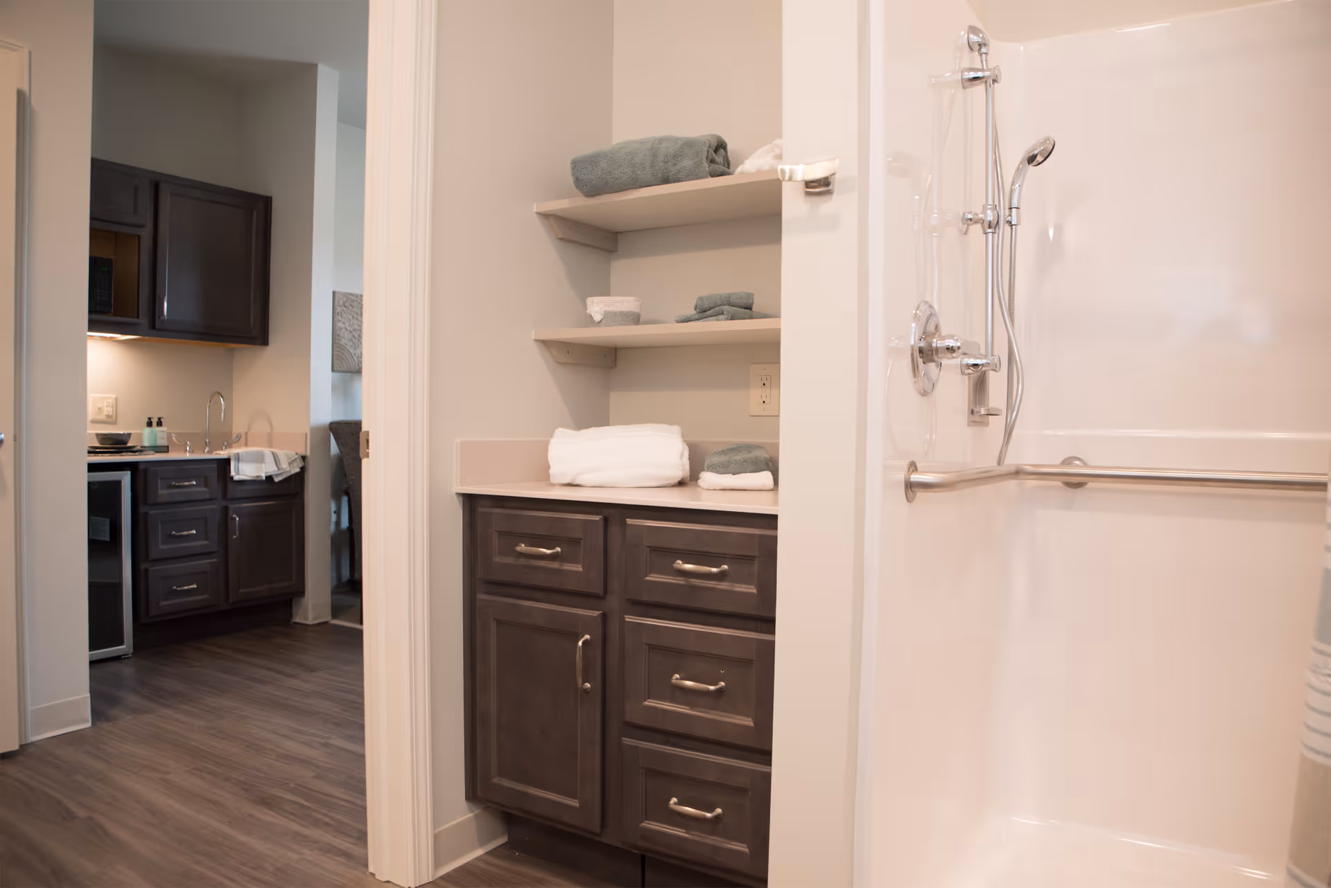 Interior view showing a bathroom area with a white shower equipped with a handheld showerhead and grab bar. Adjacent to the shower is a countertop with dark wood cabinets and open shelves holding folded towels. In the background, a kitchen area with dark cabinetry and a sink is visible.