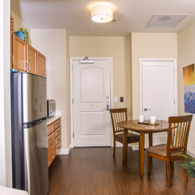 A small dining area with a round wooden table and two wooden chairs with cushions. On the table are two white mugs and a small tray. To the left is a kitchen area with wooden cabinets, a stainless steel refrigerator, and a microwave. The floor is dark wood, and the walls are light beige. There is a white door in the center background and another white door to the right. A colorful abstract painting is partially visible on the right wall.