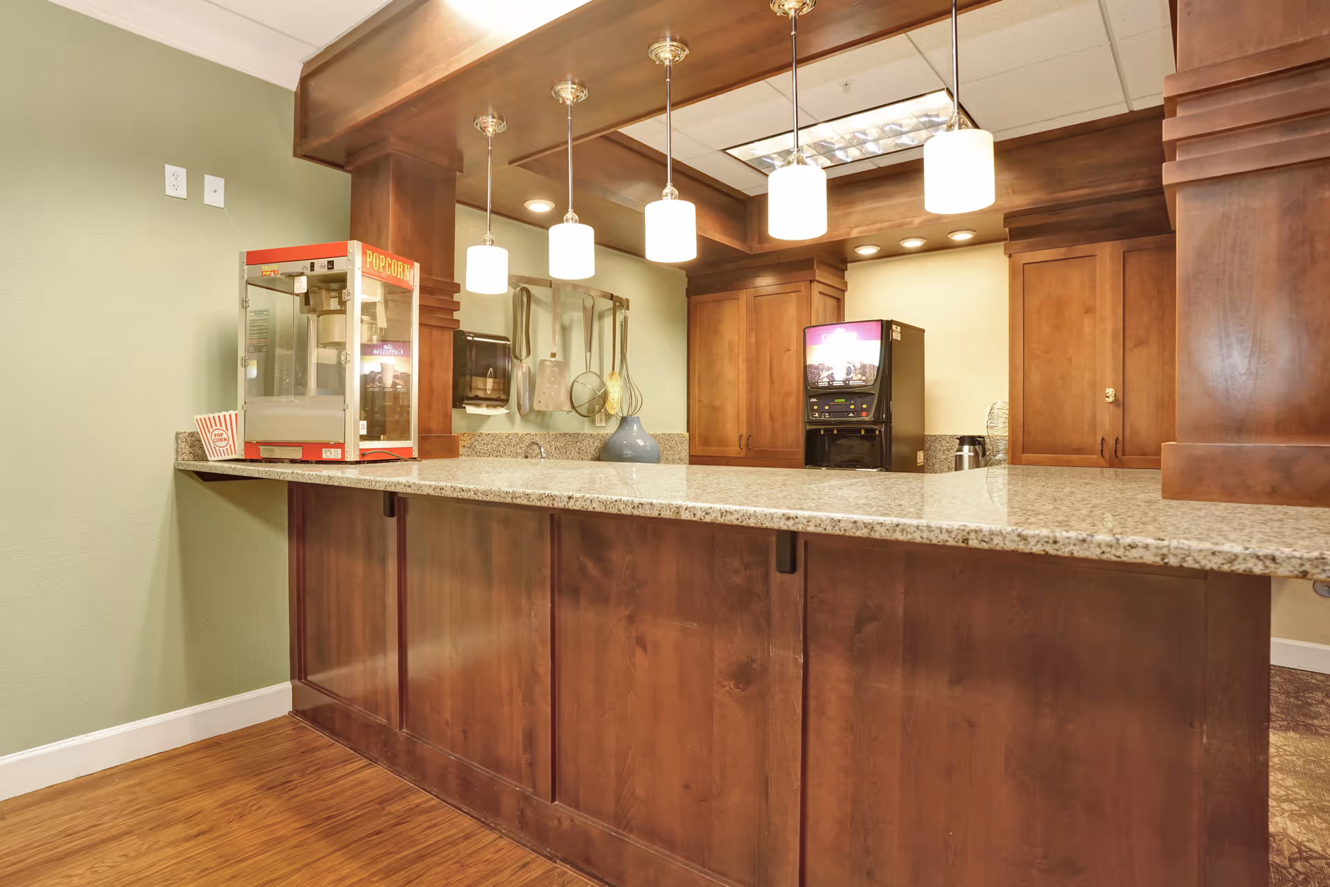Interior view of a kitchen or snack area with a granite countertop bar, wooden cabinets, hanging pendant lights, a popcorn machine on the counter, and a beverage dispenser in the background.
