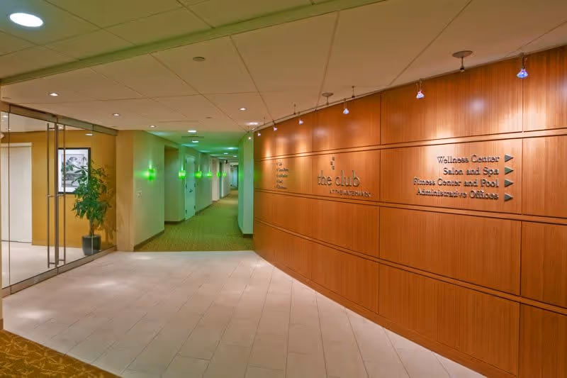 Interior hallway of a senior living facility with a wooden wall on the right displaying directional signs for the Wellness Center, Salon and Spa, Fitness Center and Pool, and Administrative Offices. The hallway is well-lit with ceiling lights and green wall sconces, and there is a glass wall with a potted plant on the left.